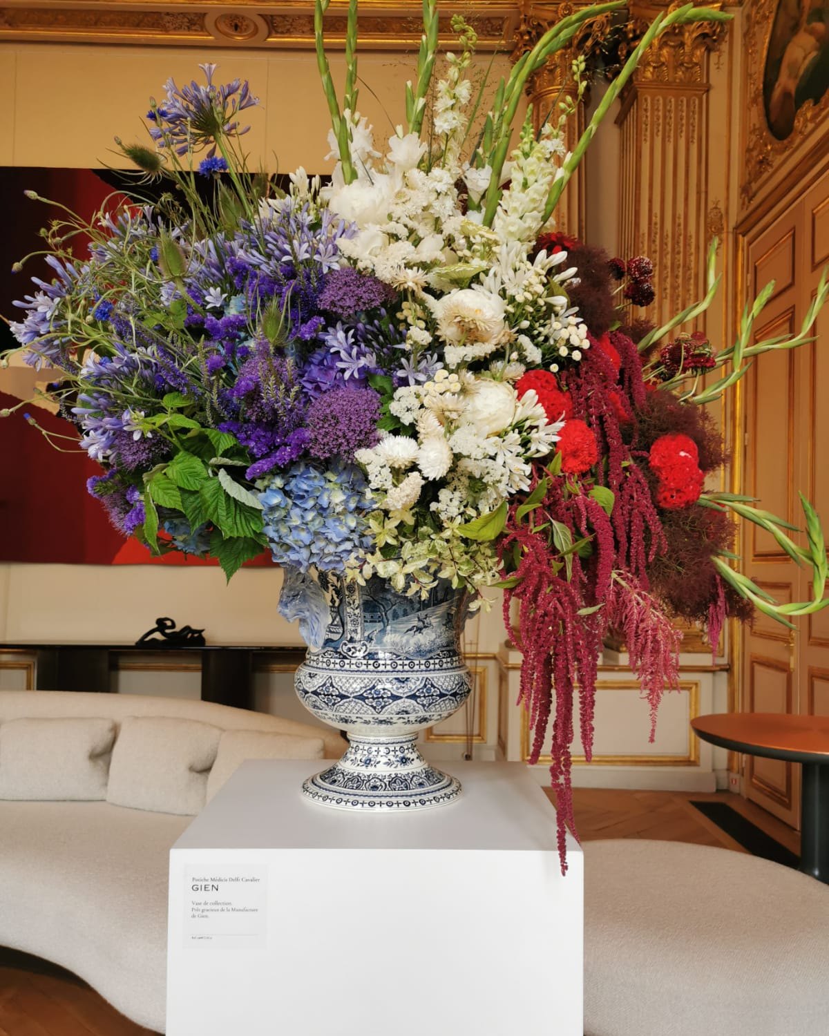 Grand bouquet bleu, blanc, rouge de fleurs françaises dans un vase Médicis en faïence de Gien, posé sur un socle blanc dans un salon doré de l'Élysée